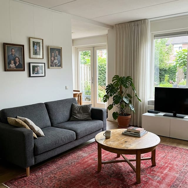 Foto van de woonkamer: grijze bank, rood tapijt, ronde houten tafel, televisie naast het raam